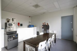 Kantineruimte met koffiemachine, houten tafel met witte stoelen en een kartonnen figuur van drie personen aan de wand.